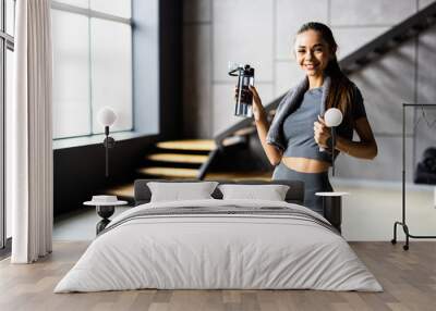 Woman drinking water at home, taking break after exercise. Smiling young attractive fitness woman with towel after training hold bottle. Wall mural